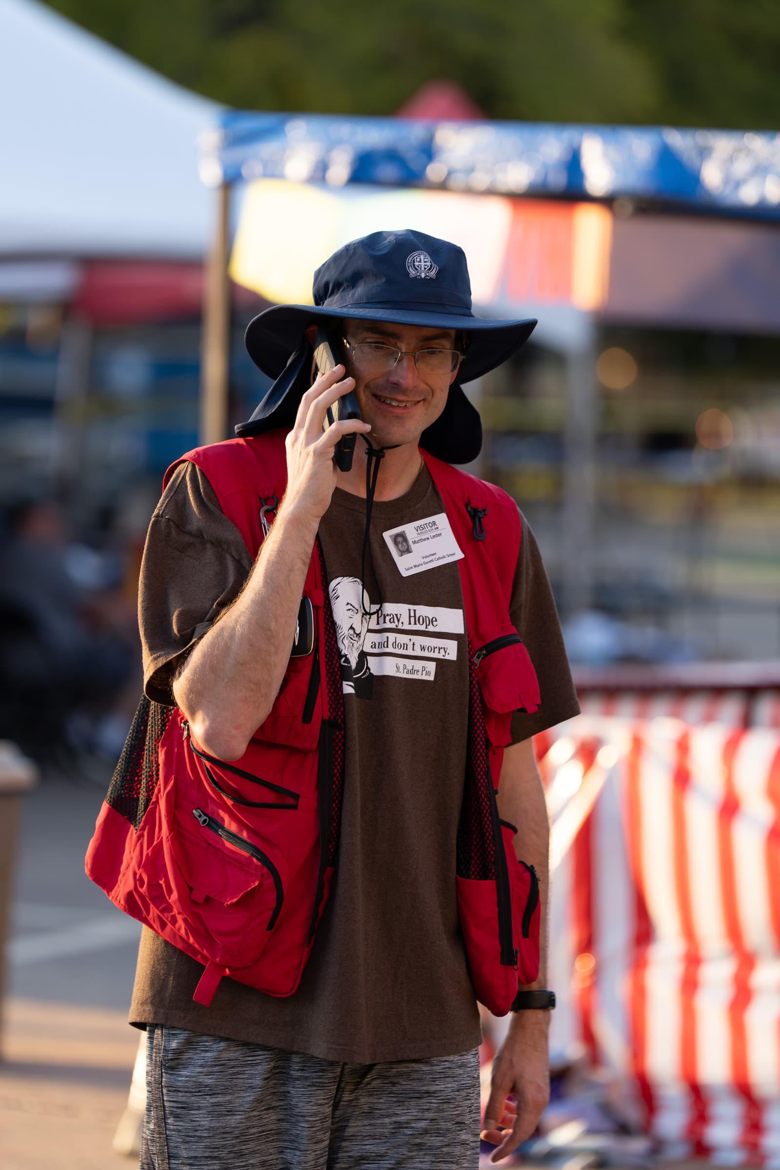 Man wearing a wide-brim hat and red vest talking on a phone while standing in front of outdoor event booths and tents.